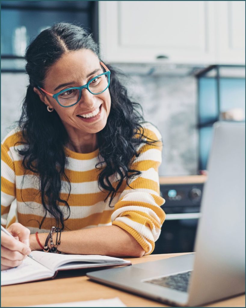 Woman with long hair and green glasses wears a yellow and white sweater and sits in front of a laptop.