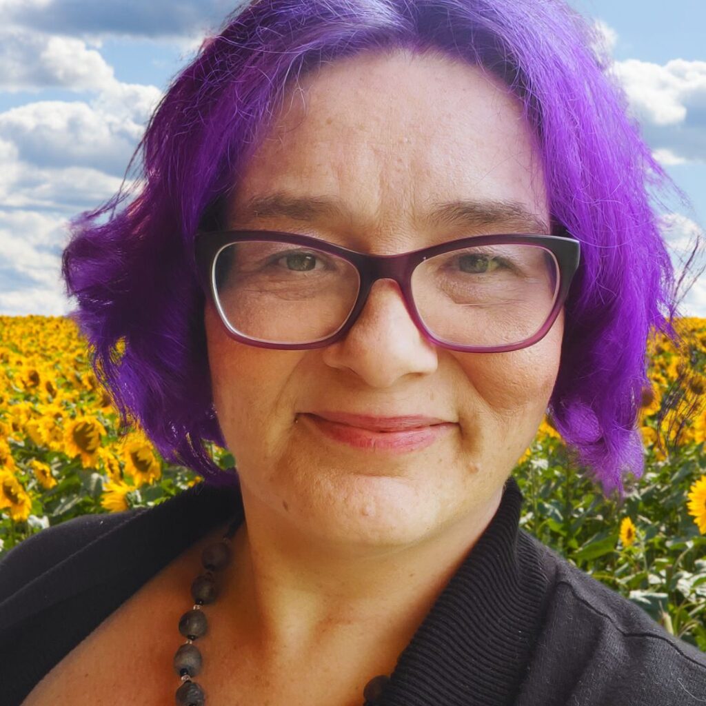 Kat, a woman with purple hair and glasses wears a black necklace and stands in front of sunflowers.