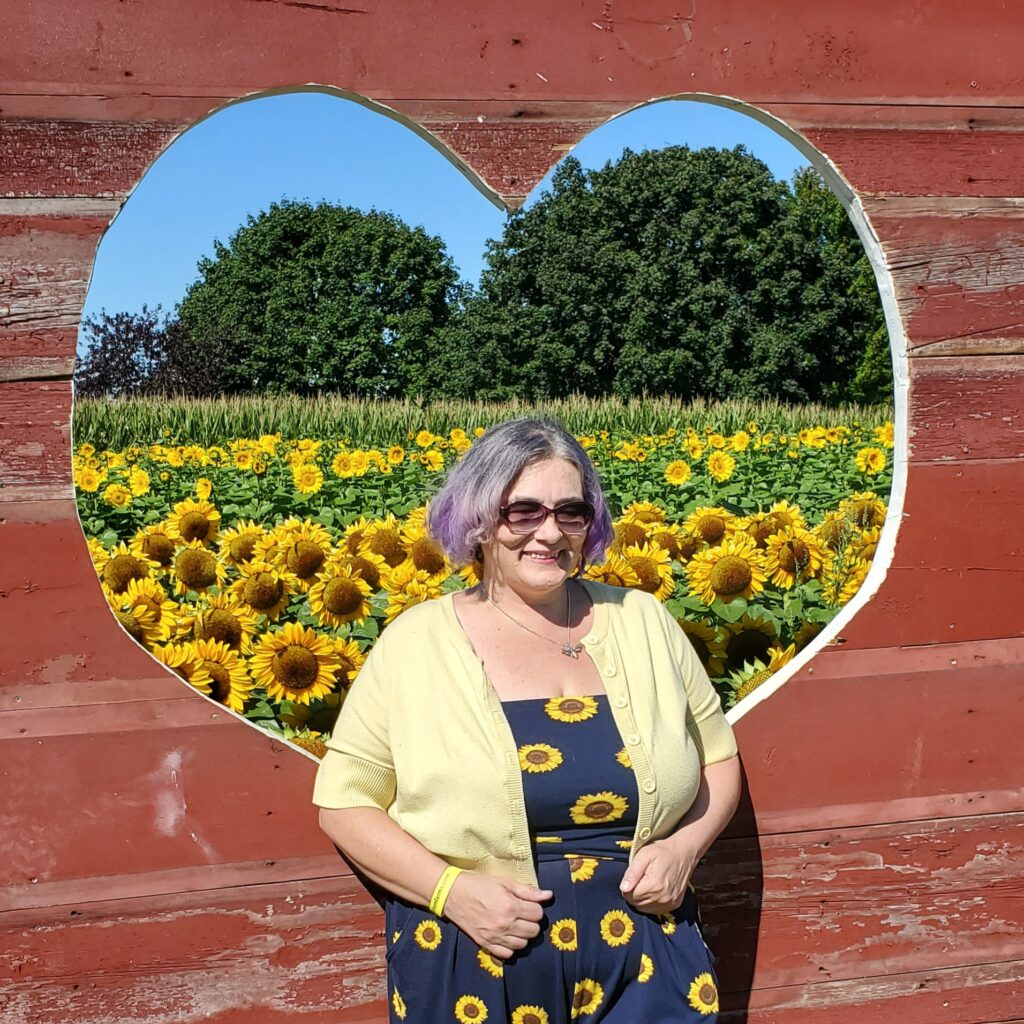 Kat, a woman with purple and gray hair is wearing a blue and yellow sunflower jumper and standing in front of a red wooden heart frame with beauitufl sunflower fields behind it.